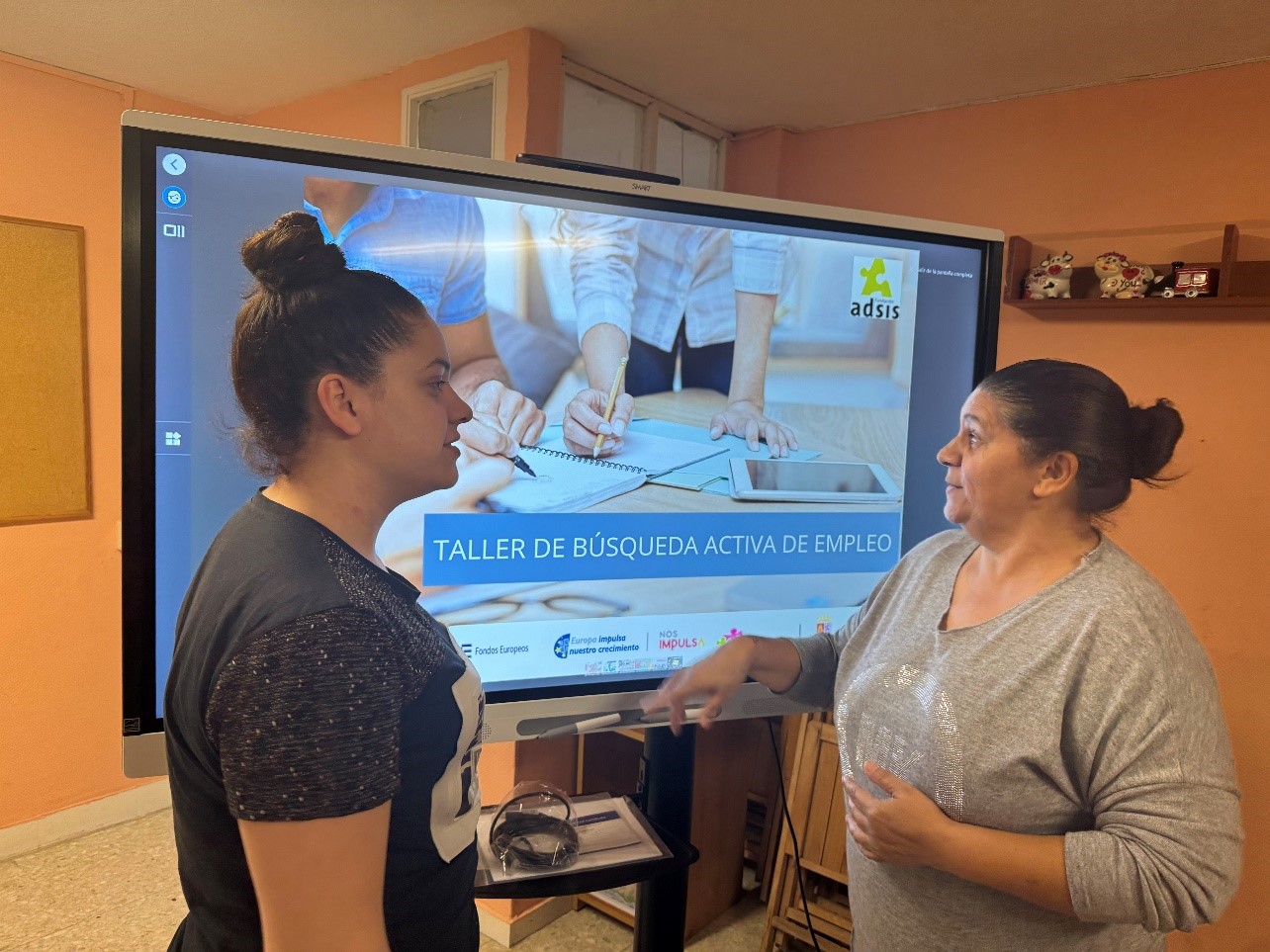 Dos mujeres hablando. De fondo una pantalla en la que se lee: Taller de búsqueda activa de empleo