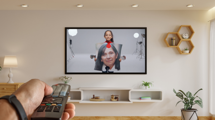 Un salón de una casa con un televisor donde se ve una de las imágenes de la campaña ContigoTodoEncaja. En primer plano, una mano sujetando un mando de tv.