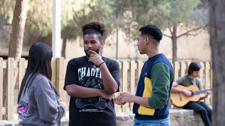 tres jóvenes charlando en un parque, en el fondo, otro toca la guitarra