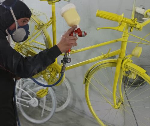 Un chico con mascarilla y traje de protección pintando una bici de color amarillo con spray