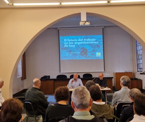 Público sentado en una sala de actos desde atrás. Al fondo, una pantalla con una presentación