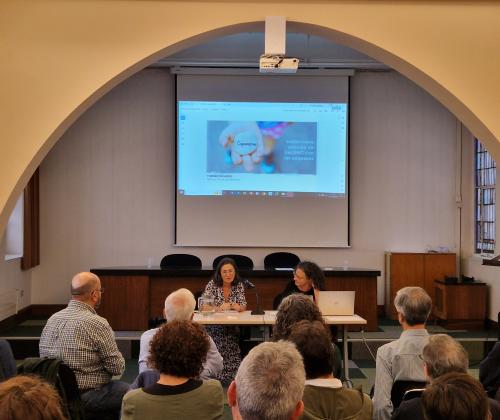 Público sentado en una sala de actos desde atrás. Al fondo, una pantalla con una presentación