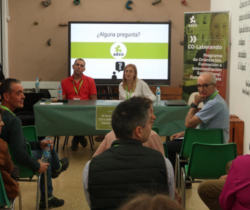 Dos personas sentadas en una mesa con una pantalla de fondo con el logo de Fundación Adsis