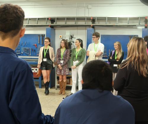 De espaldas tres jóvenes con uniforme de taller hablando con un grupo de personas